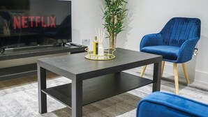 A modern living room with a flat-screen TV displaying 'NETFLIX', a dark wooden coffee table, a blue armchair, and a small plant on the table. - Modern 1BR Flat | Walkable High Street Stay (Southend-on-Sea)