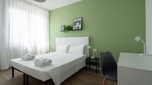 A neatly made bed with white linens and towels, a bedside table with a lamp, a desk with a chair, and a green wall with framed pictures. - Carducci Modern Apartment 7 by Wonderful Italy (Mestre)
