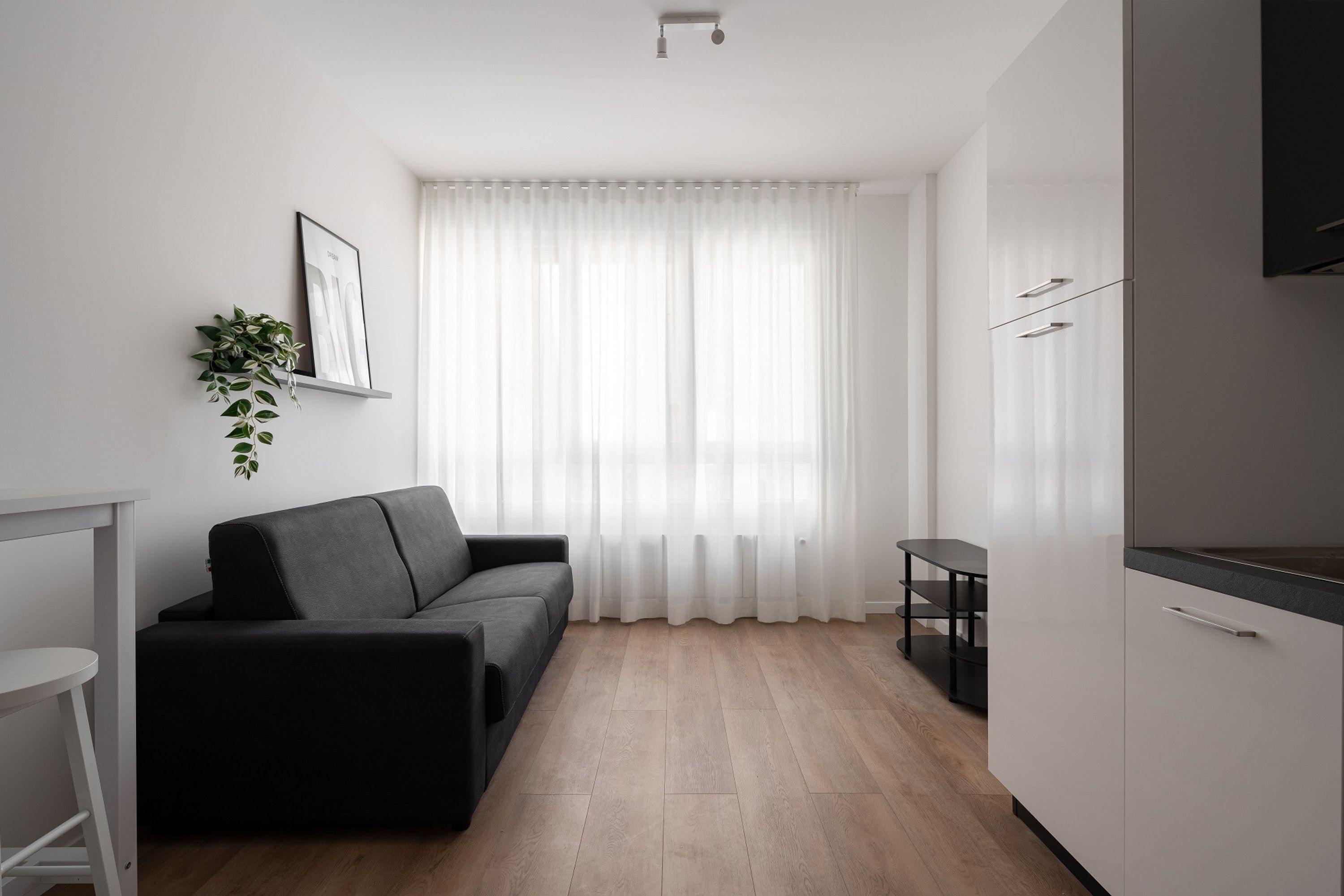 A modern, minimalistic living space with a black sofa, a small white table, and a kitchen area with white cabinets and a black countertop.