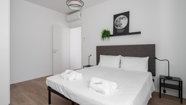 Egy modern hálószoba, melyben egy nagy ágy, fehér ágynemű, sötét színű ágyvég, egy éjjeliszekrény, a falra szerelt légkondicionáló, egy keretezett holdfényképet ábrázoló kép és egy kis növény található.