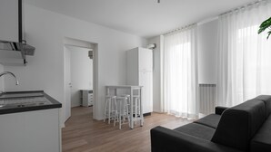 Egy modern konyha fehér szekrényekkel és fekete munkalappal, egy étkező rész fehér bárszékekkel, és egy fekete kanapé egy világos nappaliban, ahol áttetsző függönyök díszelnek.