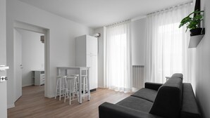 Egy modern nappali, melyben egy fekete kanapé, egy fehér étkező bárszékekkel, és a háttérben egy konyha található.
