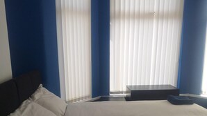 Une chambre à coucher avec un grand lit, une table de chevet et une chaise. Il y a deux fenêtres équipées de stores.
