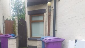 Une ruelle étroite avec deux poubelles violettes et une poubelle bleue, un bâtiment blanc avec une fenêtre et une porte, et un coffret électrique blanc sur la droite.