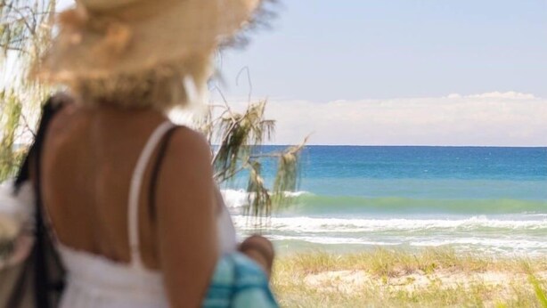 Una persona con sombrero de paja contemplando una playa con olas y palmeras.