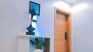 A hallway with a wooden door, a white shelf with a potted plant, and a wall-mounted lamp. - Klulead Garden and Suites (Ijebu Ode)