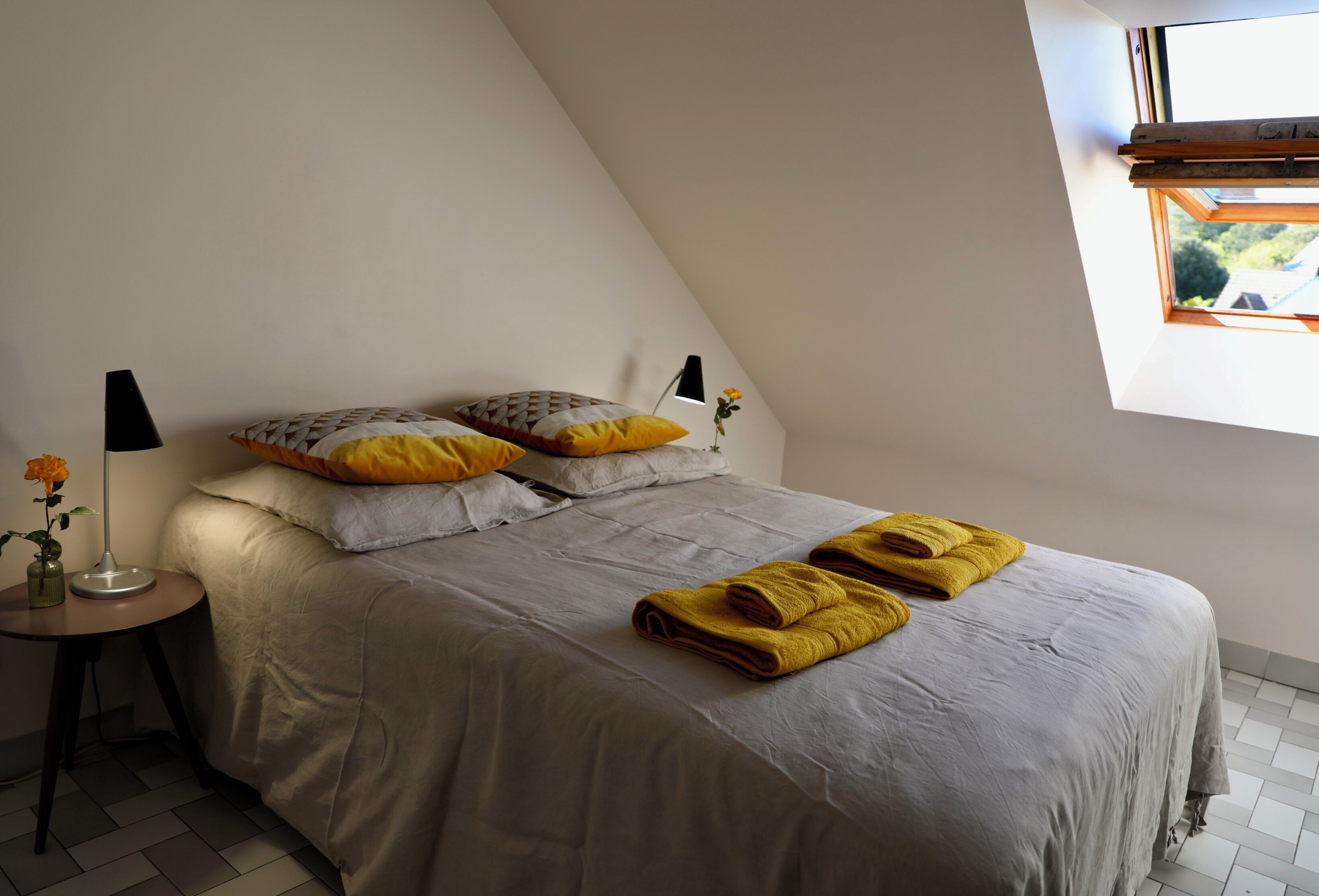 Een slaapkamer met een bed, kussens, een nachtkastje met lamp en een raam met uitzicht.