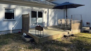 A patio area with a wooden deck, a table with chairs, a large umbrella, and a sliding glass door leading to a covered porch. - In the Middle of the Forest Nearby Ahus (Aahus)