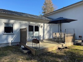 Un espace patio avec une terrasse en bois, une table entourée de chaises, un grand parasol et une porte-fenêtre coulissante donnant sur un porche abrité.