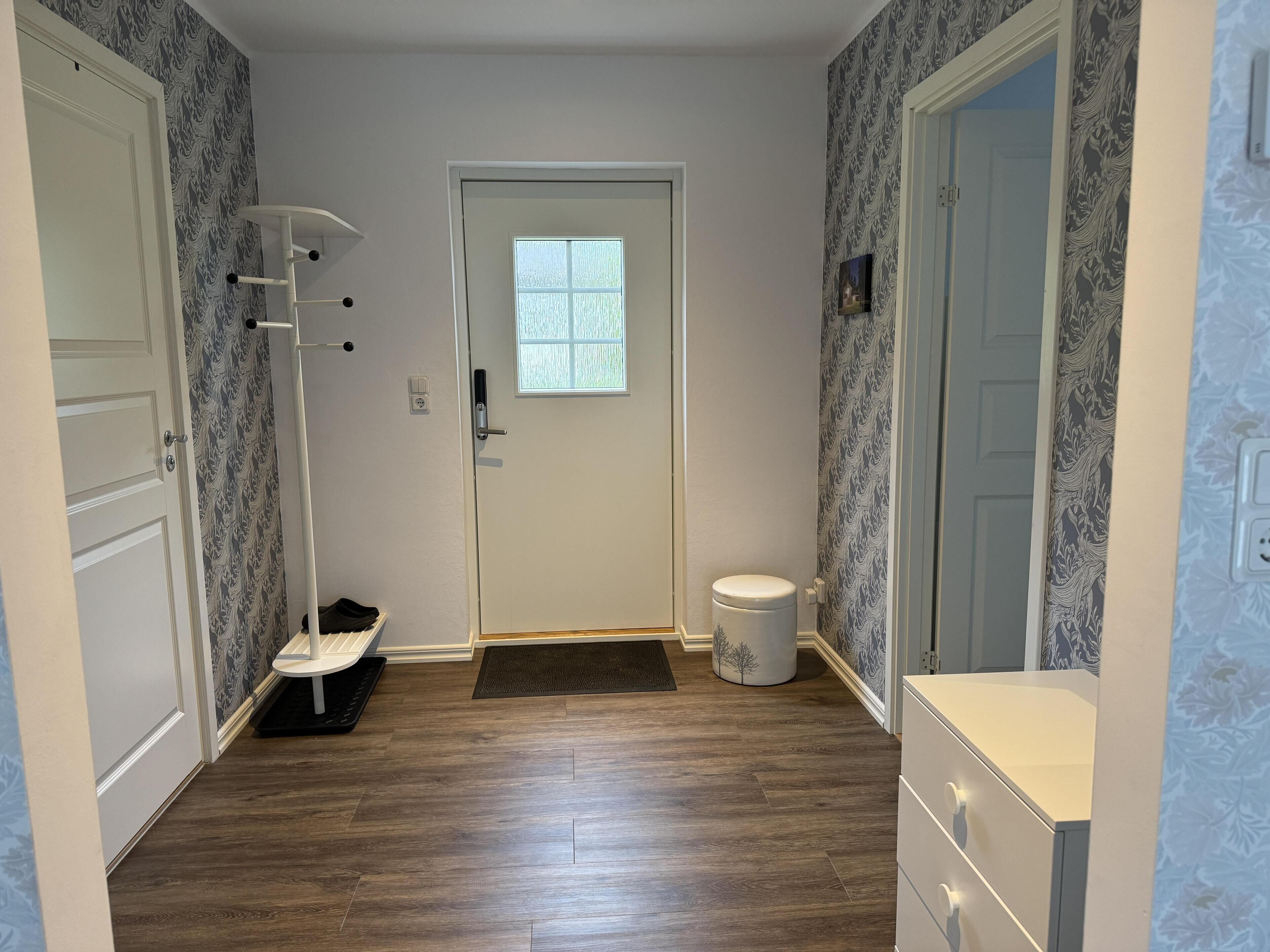 A narrow hallway with a white door, a small window, a white bench, and a trash can.