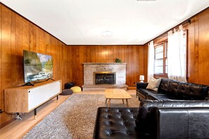 A living room with wooden walls, a stone fireplace, a black leather sofa, a wooden coffee table, and a television on a wooden stand.