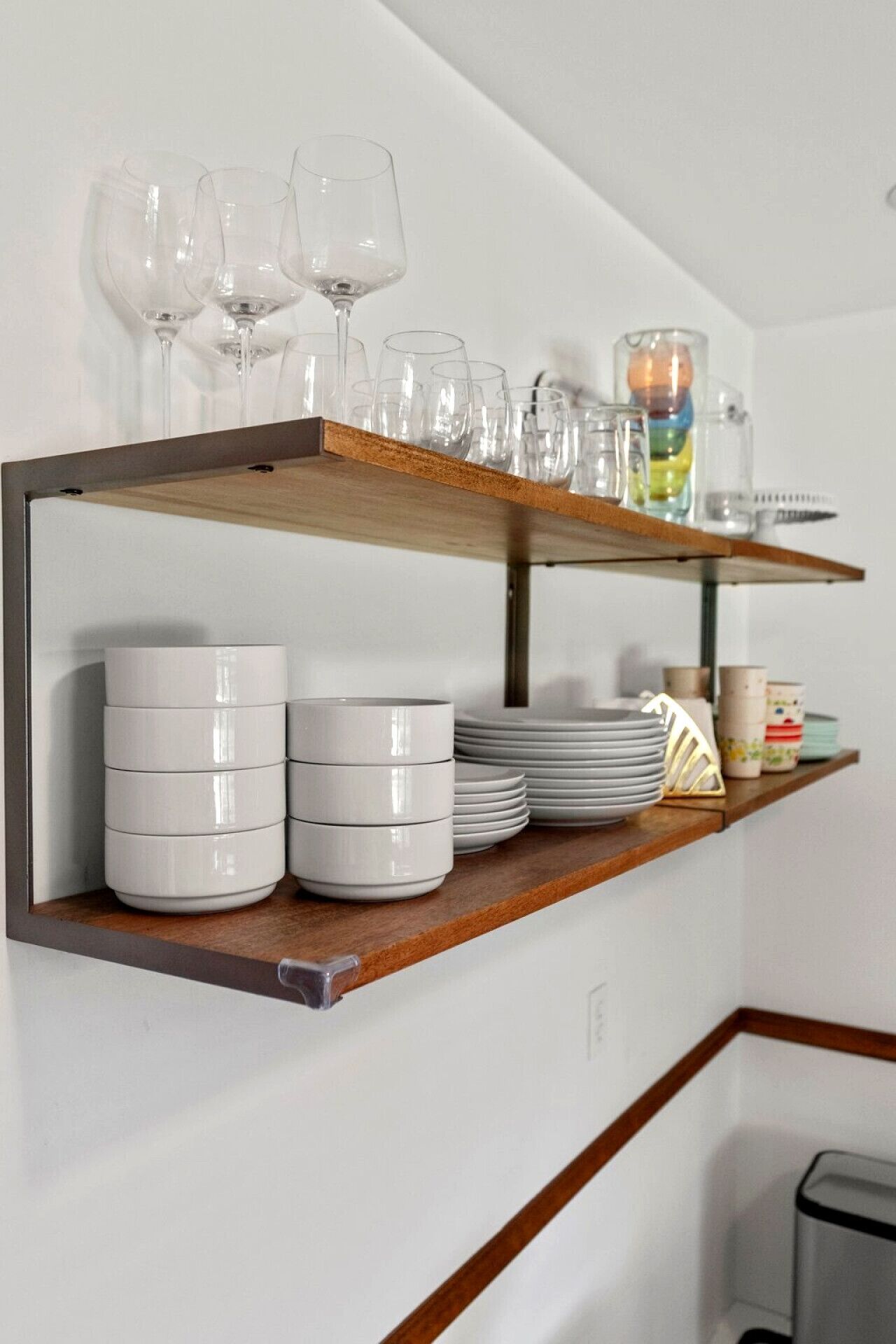 A kitchen shelf with stacked white plates, a stack of white bowls, and a row of clear wine glasses.
