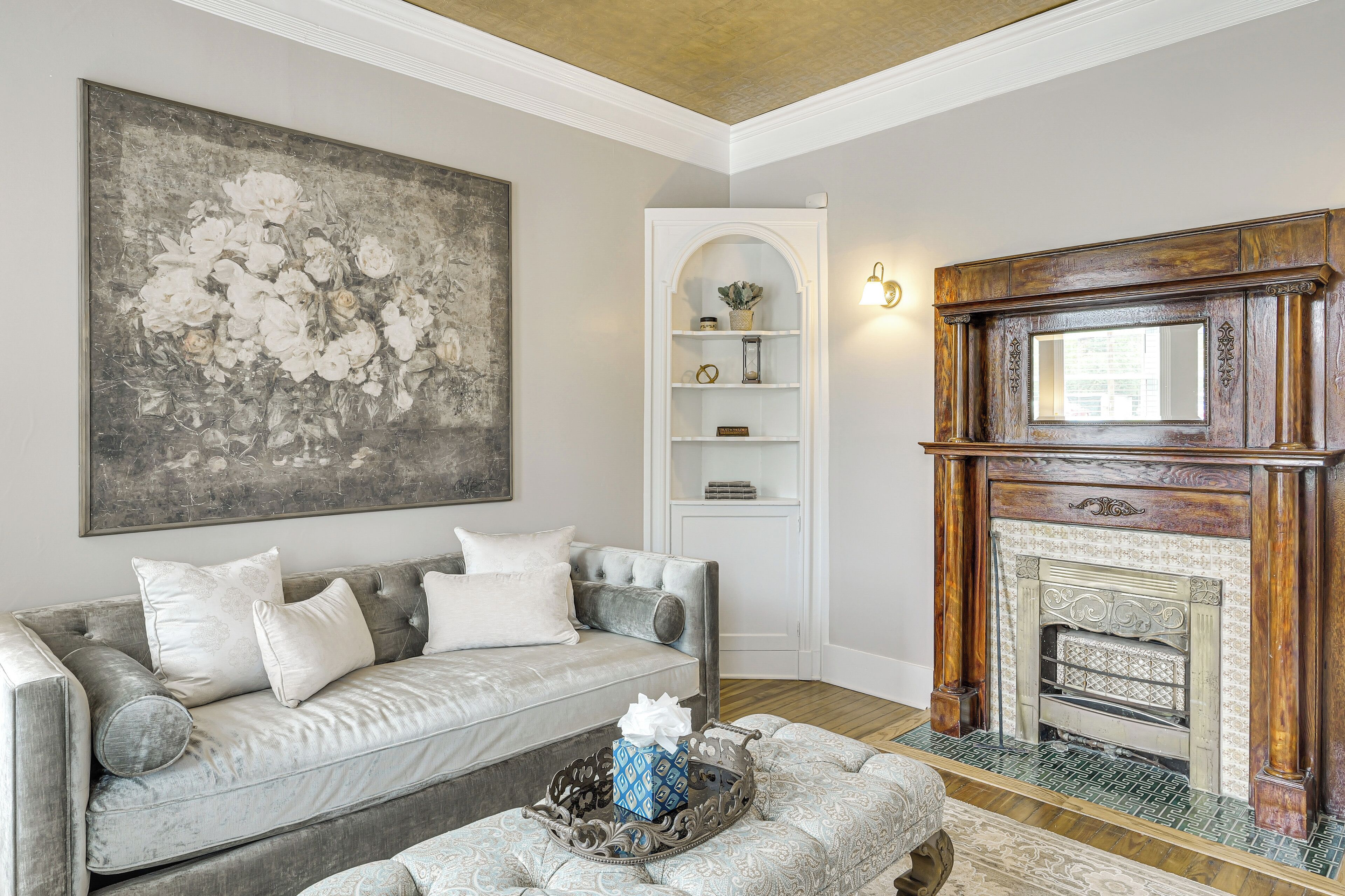 Un salon avec un canapé gris, une cheminée et un grand tableau représentant des motifs floraux.