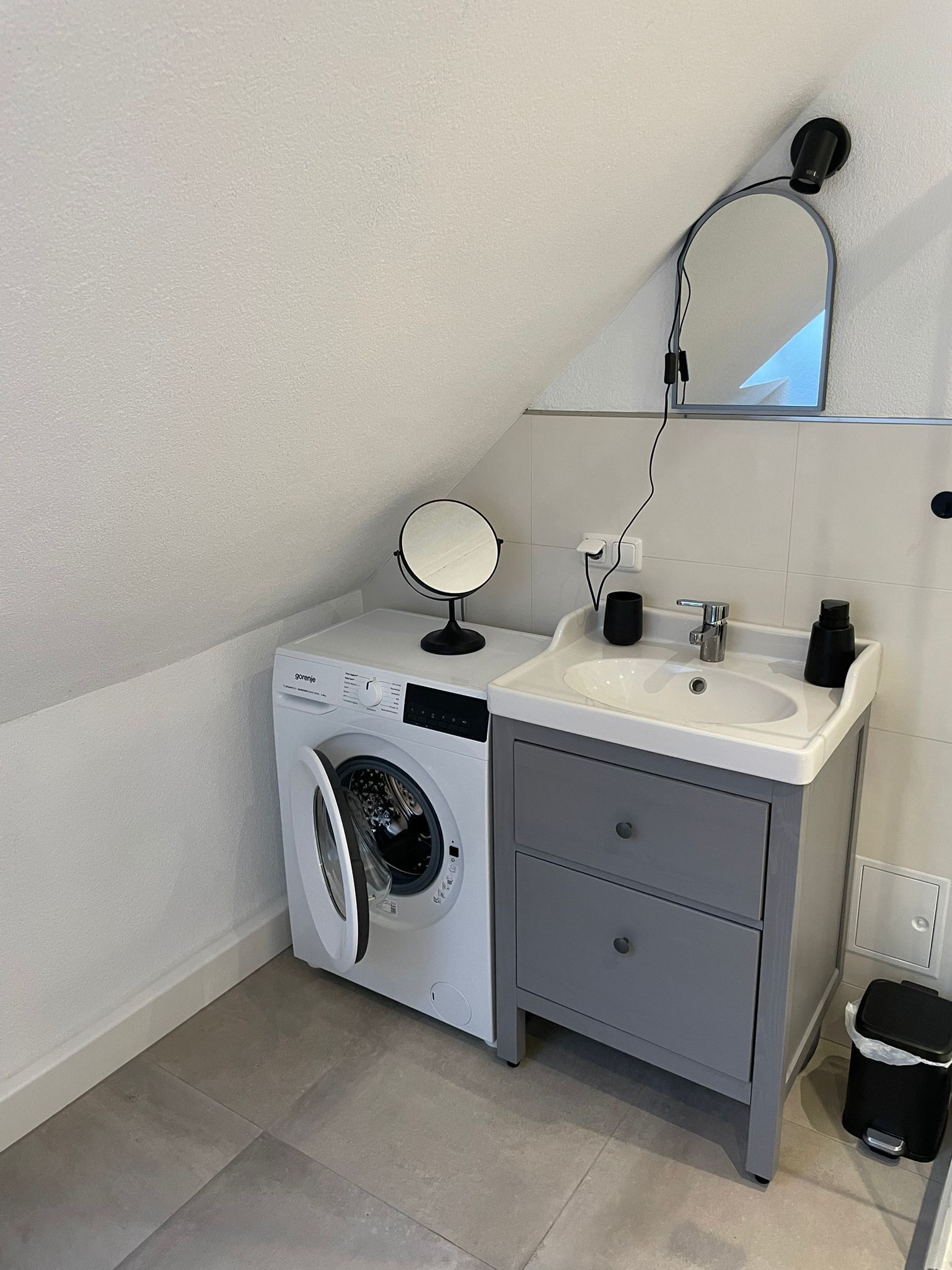 Un baño pequeño con lavadora, lavamanos, espejo y cubo de basura.