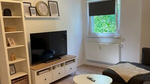 Una sala moderna con un televisor de pantalla plana sobre un mueble de madera, un aparador blanco con objetos decorativos, una mesa de centro redonda y un sofá de color oscuro.
