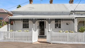 A white two-story house with a gabled roof, a front porch with decorative ironwork, and a white picket fence.