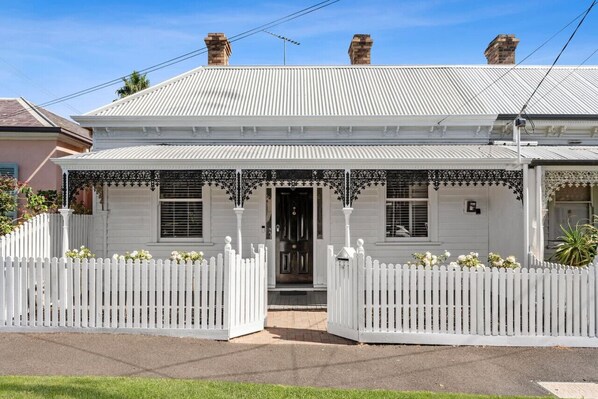 A white two-story house with a gabled roof, a front porch with decorative ironwork, and a white picket fence.
