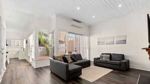 A modern living room with dark wood flooring, two black sofas, a beige rug, and a kitchen in the background.