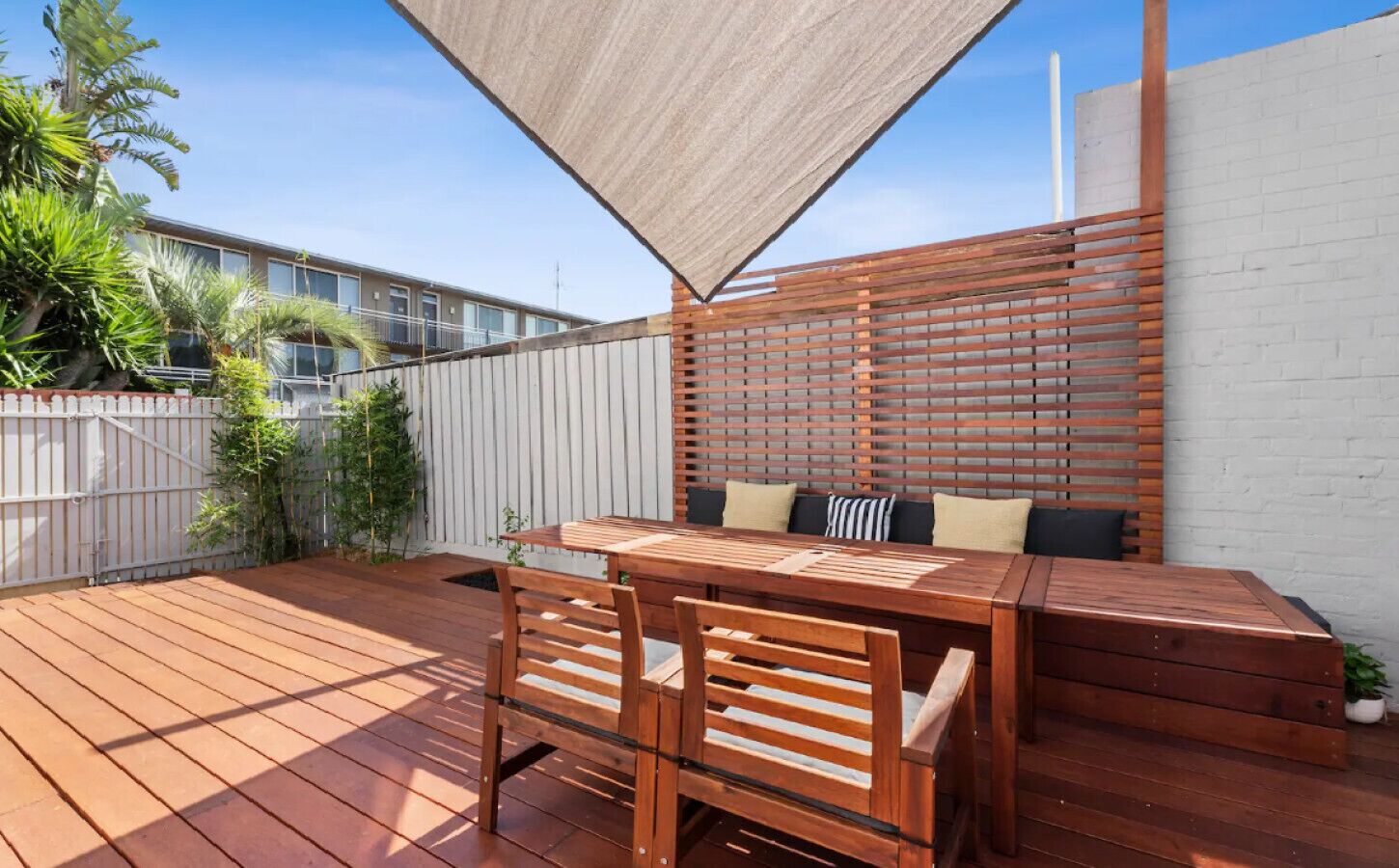 Un patio al aire libre con una mesa y sillas de madera, una zona de estar con cojines y una cerca blanca.