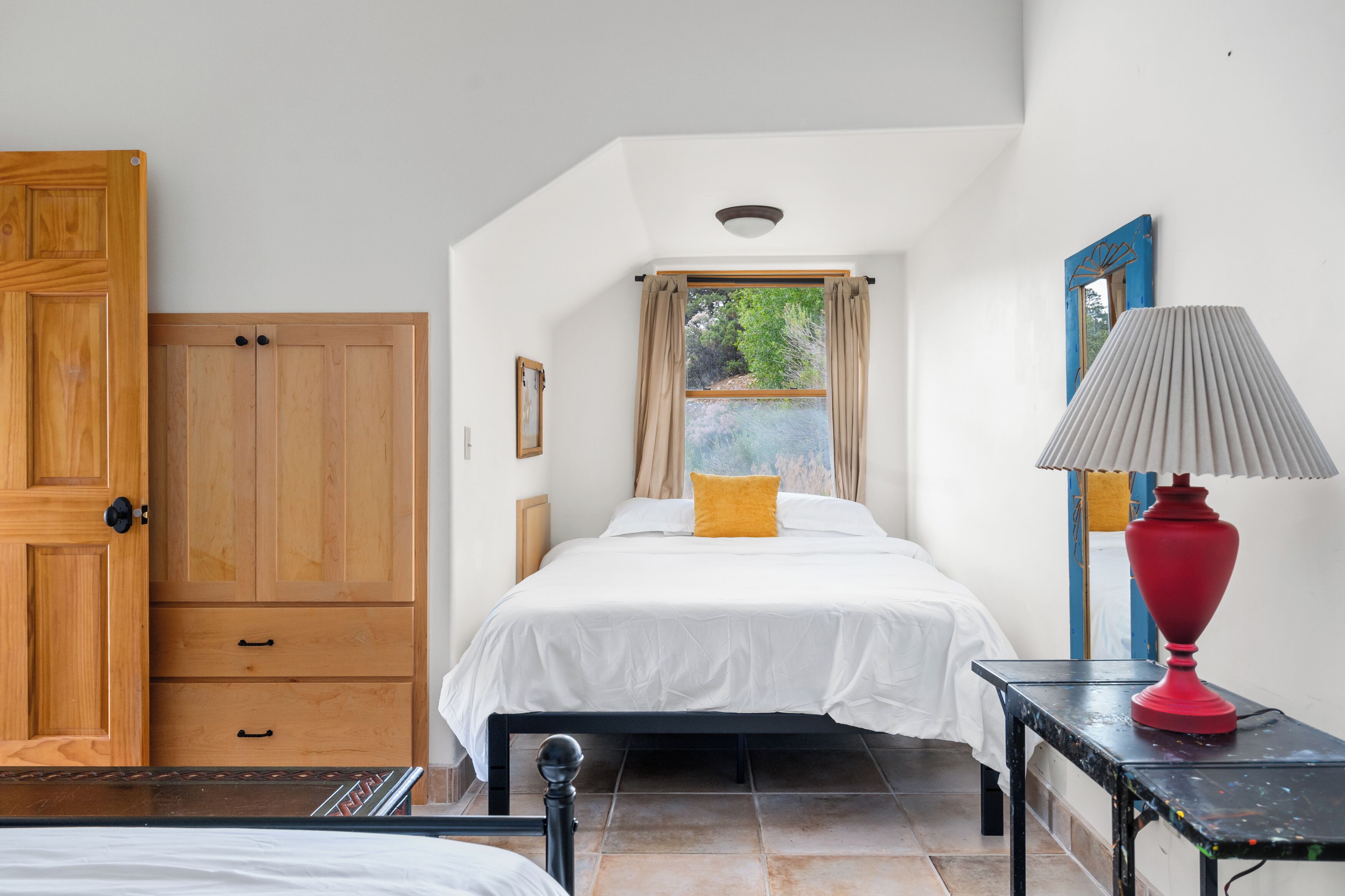 Een slaapkamer met een bed, nachtkastje, rode lamp, raam met gordijnen, houten garderobe en een deur.
