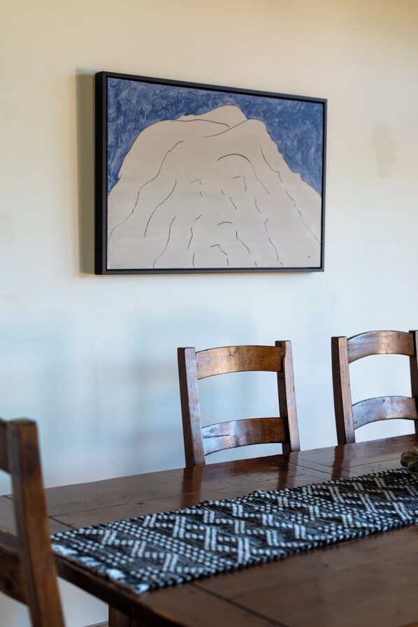 A dining table with two wooden chairs and a patterned placemat. A framed artwork depicting a mountain is hung above the table.