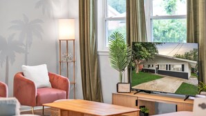 A living room with a red armchair, a wooden coffee table, a TV on a stand, a floor lamp, and a view of a house through a window. - Charming 3bd - Near beaches - Foosball & Backyard (Norfolk)