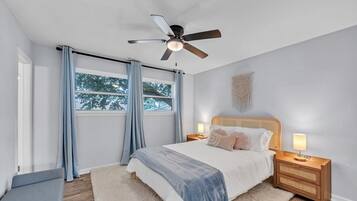 A bedroom with a bed, nightstands, a ceiling fan, a window with curtains, and a wall-mounted macrame piece.