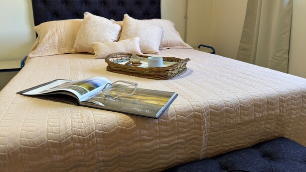 A neatly made bed with a quilted bedspread, a dark headboard, and several pillows. A wicker tray with a cup and cookies is placed on the bed. - Departamento cerca del centro (El Calafate)