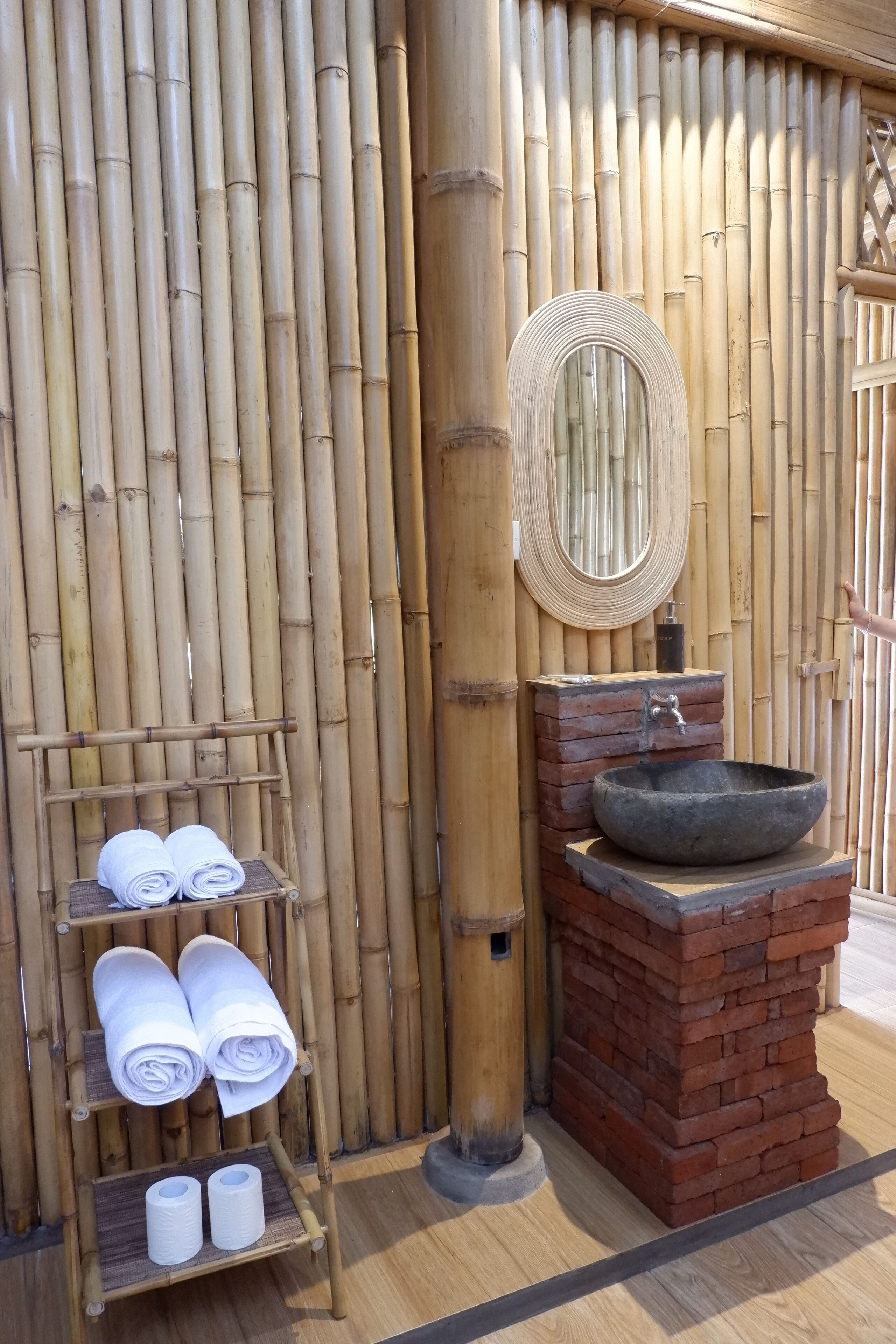 Un mur en bambou agrémenté d’un miroir rond et d’un lavabo en pierre. Des serviettes et du papier toilette sont posés sur une étagère.