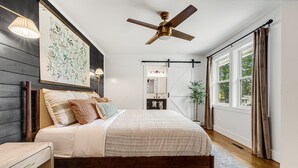 A bedroom with a wooden bed, a ceiling fan, a large window with curtains, a nightstand, and a wall-mounted mirror. - Quiet Furnished 3BR Home Near Asheville - Fast WiFi, Firepit (Canton)
