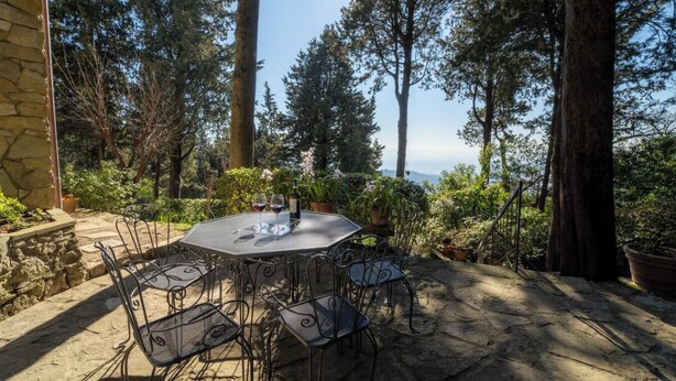 Eine Terrasse im Freien mit einem runden Tisch und Metallstühlen, umgeben von hohen Bäumen und einer Steinmauer.
