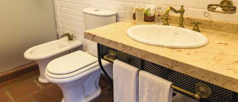 Un baño con inodoro, bidé y lavabo con encimera de mármol. Hay artículos de aseo en el lavabo y un perchero con toallas.