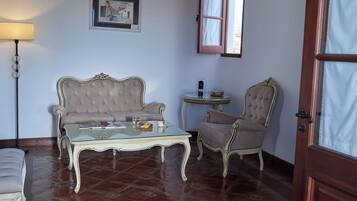 Una habitación con un sofá capitonado, una mesa de centro de vidrio, una mesita auxiliar, una silla, una lámpara y un cuadro en la pared.