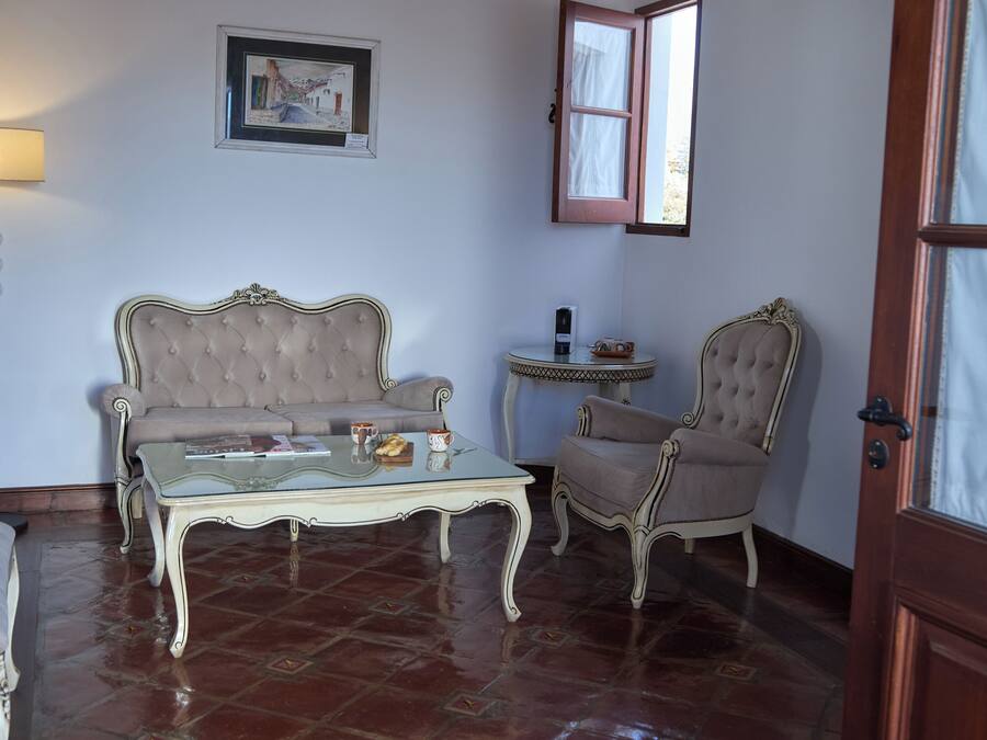 Una habitación con un sofá capitonado, una mesa de centro de vidrio, una mesita auxiliar, una silla, una lámpara y un cuadro en la pared.