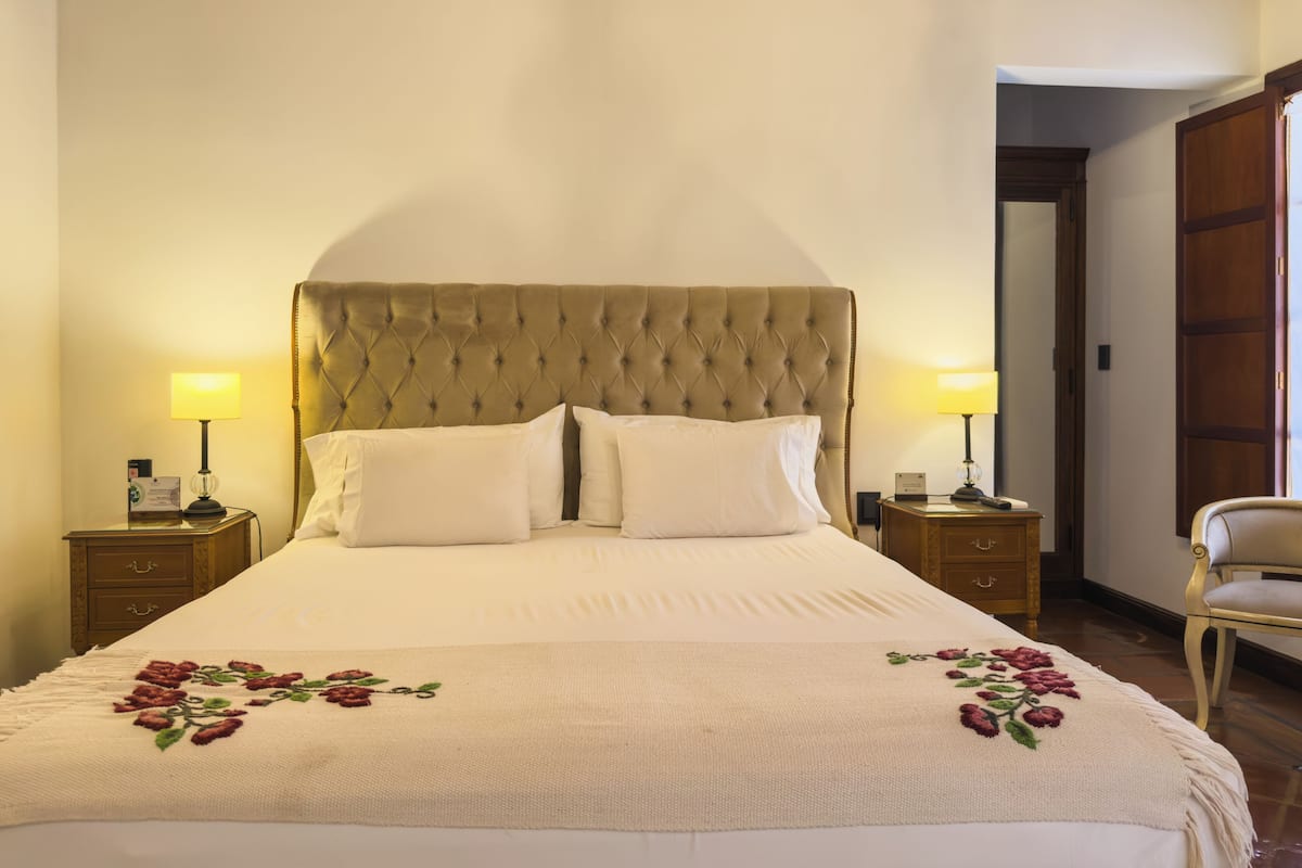 Una cama bien tendida con cabecera capitonada, dos mesitas de noche con lámparas y un plaid estampado de flores.