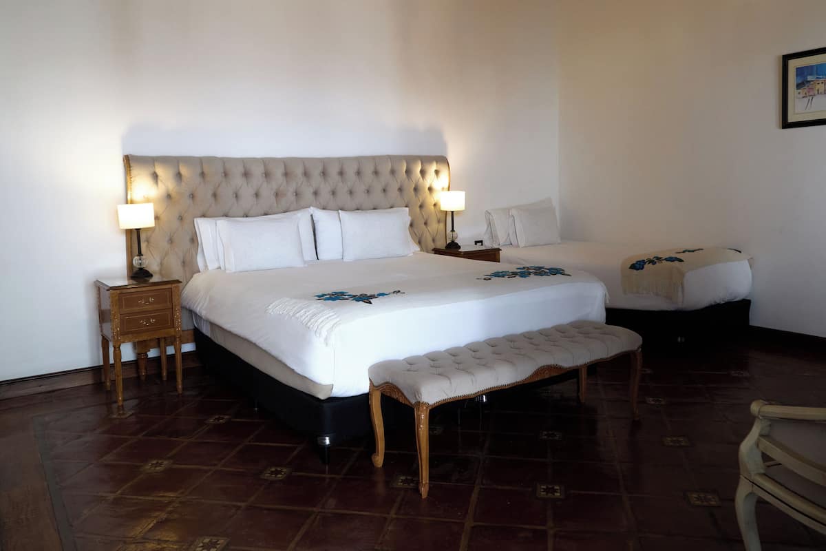 Una cama doble con cabecera capitonada, dos mesitas de noche con lámparas, un banco y una silla de madera.