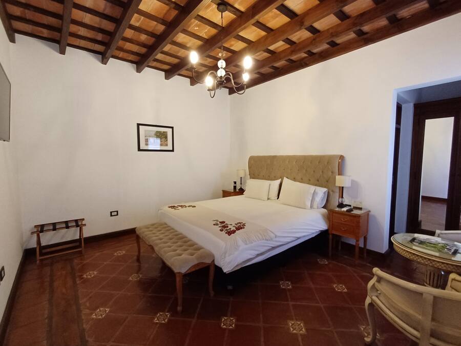 Un dormitorio con una cama grande, cabecera de madera, lámparas de noche, un banco y una pequeña mesa con una silla.