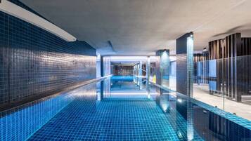 Une piscine intérieure moderne, dotée d’un sol et de murs recouverts de carreaux bleus, entourée de rampes de verre et bordée d’un couloir dont le plafond est également recouvert de carreaux.