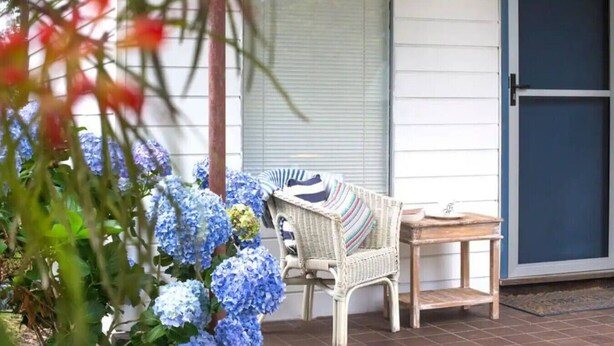 A porch with a wicker chair and a small table, a blue door, and a garden with hydrangeas. - Cosy retro styled bungalow (Mollymook Beach)