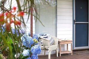 A porch with a wicker chair and a small table, a blue door, and a garden with hydrangeas. - Cosy retro styled bungalow (Mollymook Beach)