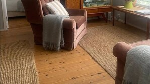 A living room with a brown armchair, a glass-top coffee table, a framed picture on the wall, and a window with blinds. - Cosy retro styled bungalow (Mollymook Beach)