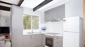 A modern kitchen with grey cabinets, a white refrigerator, a built-in oven, and a window with a view of greenery. - Cosy retro styled bungalow (Mollymook Beach)