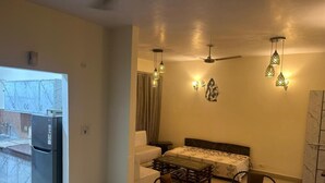 A compact bedroom with a bed, a dining table, and chairs. There are ceiling fans and wall-mounted lights. A refrigerator is visible in the adjacent room. - Highriseviews/familyfriendly/neartajmahal (Agra)