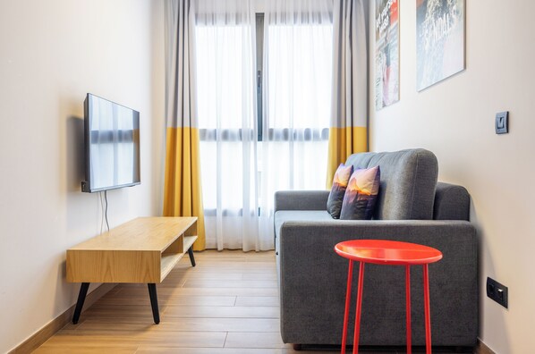 A modern living room with a grey sofa, a wooden side table, a red stool, and a flat-screen TV mounted on the wall. - Sholeo Lodges Las Palmas Flats (Las Palmas de Gran Canaria)