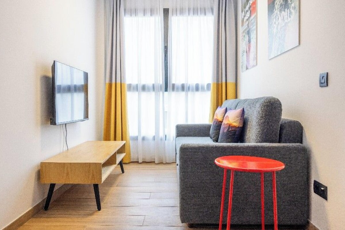 Uma sala de estar moderna com um sofá cinzento, uma mesa de centro de madeira, um banquinho vermelho e uma TV fixada na parede.