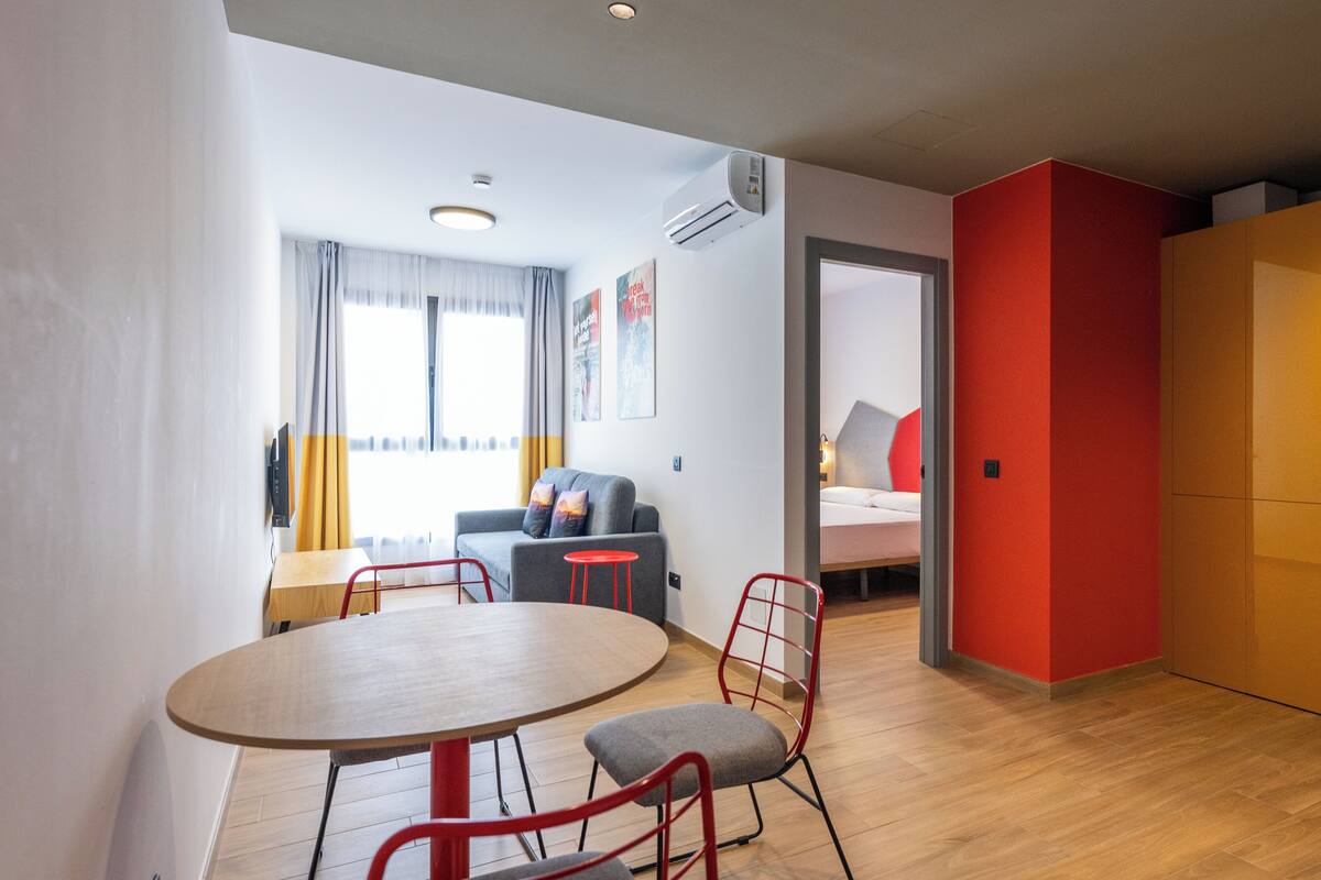 Ein moderner, kompakter Wohnraum mit Essbereich, einer Küchenzeile und einem Schlafzimmer mit roter Akzentwand.