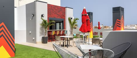 An outdoor patio with tables and chairs, a red umbrella, and a view of the city.
