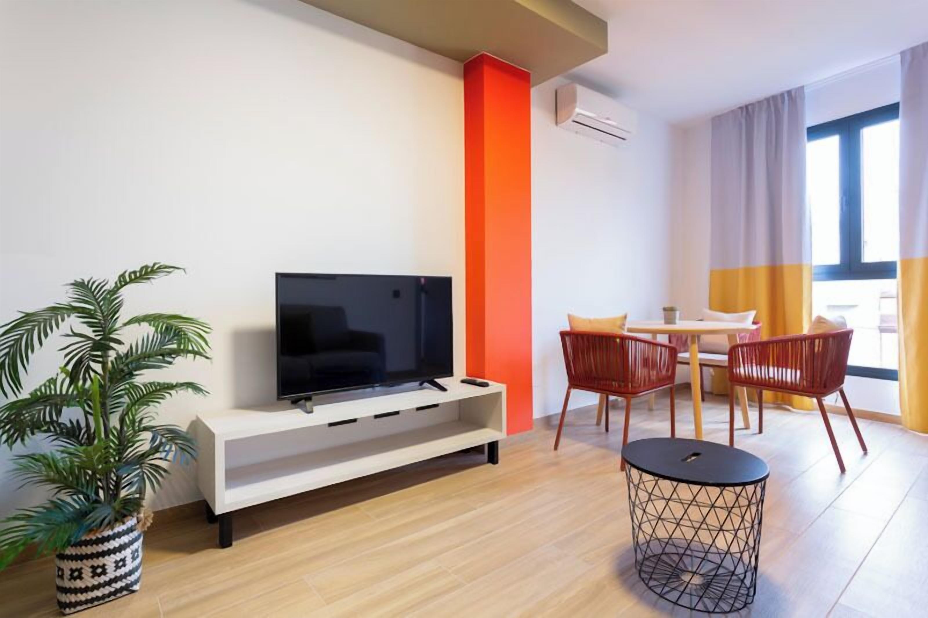 A modern living room with a flat-screen TV on a white console, a dining area with a table and chairs, a potted plant, and a round black side table.
