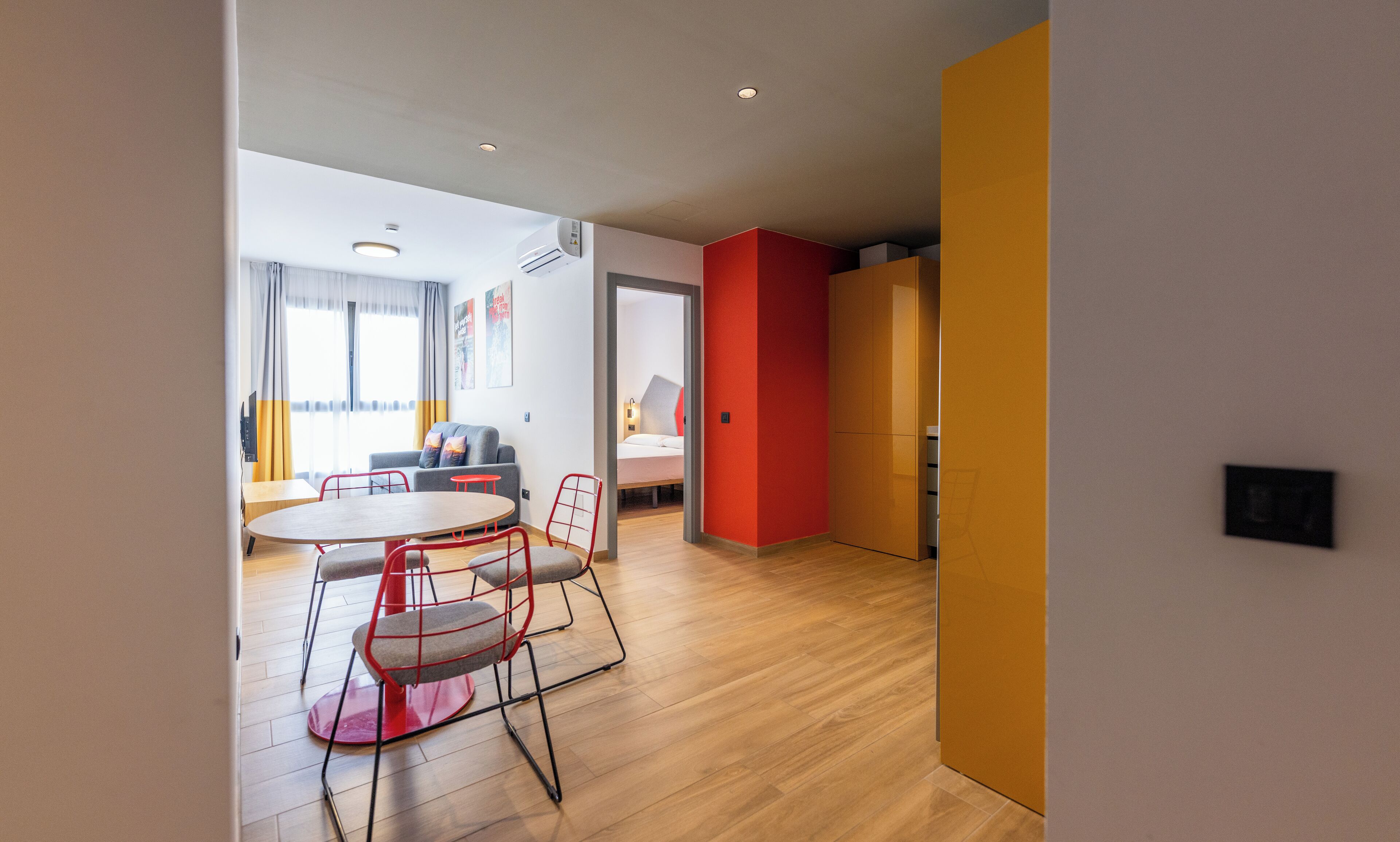 A modern living room with a round wooden table, red chairs, a grey sofa, and a red accent wall.