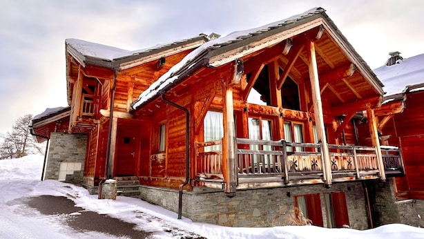 A log cabin with a stone foundation, a balcony with a metal railing, and a snow-covered roof.